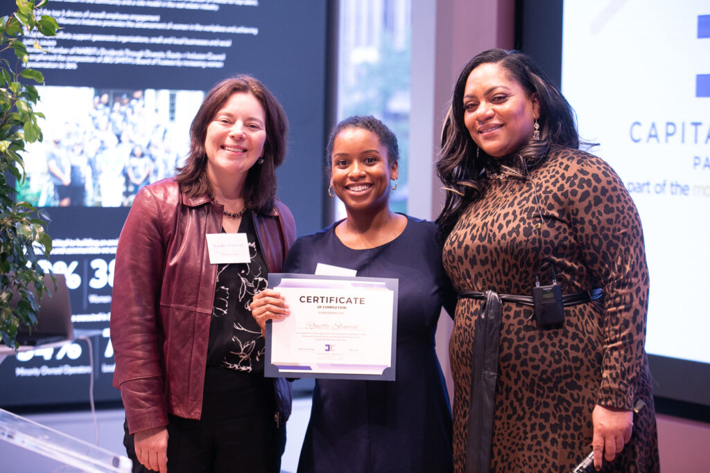 Black woman in a black dress receiving a certificate while standing between two other women