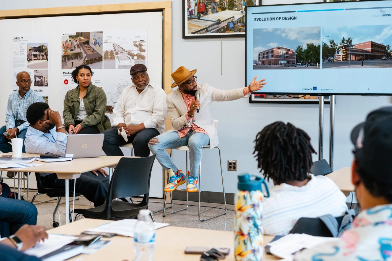 Participants in an Equitable Development Initiative cohort in Detroit sit in high chairs behind a table, making a presentation about a proposed real estate development. One person holds a microphone in his right hand and gestures with his left arm outstretched, gesturing toward a screen showing two images of the proposed building.
