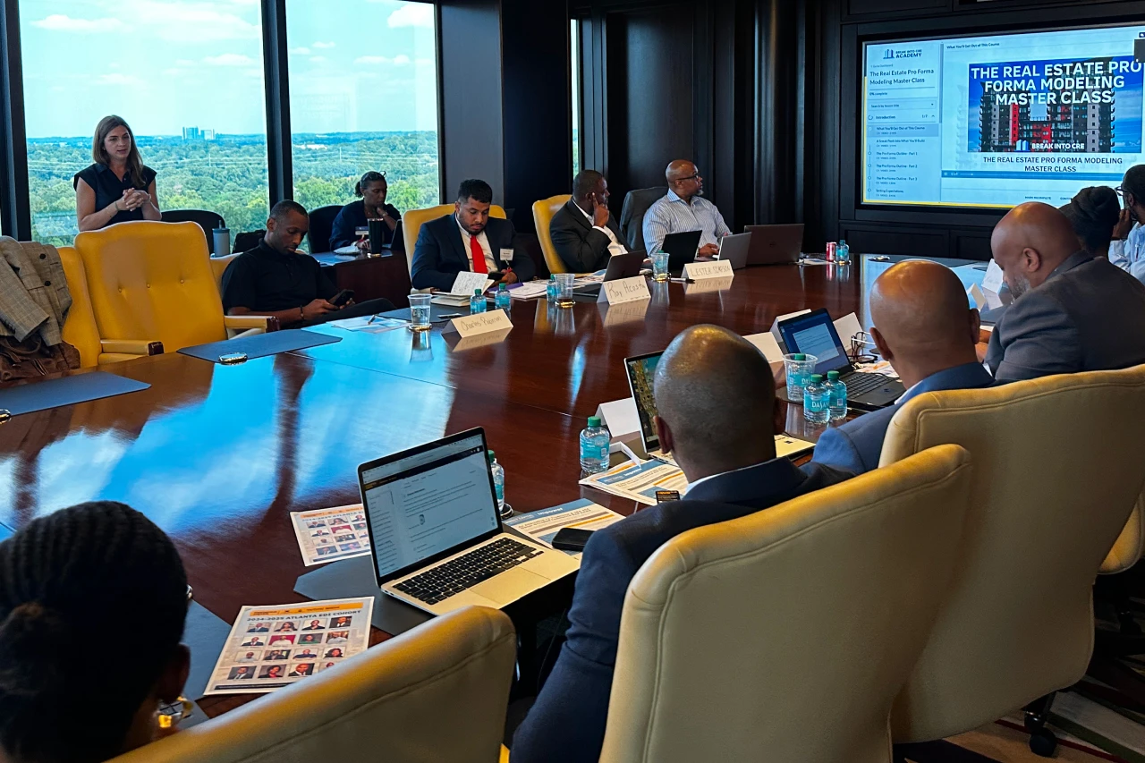 10 people sit on both sides of a long table, looking forward at a screen with a presentation titled "The Real Estate Pro Forma Modeling Master Class"