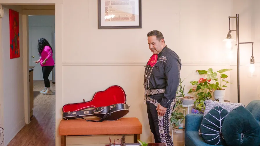 Enrique Leon, an owner of Pilsen Housing, wears a mariachi outfit and stands next to his guitar case at home